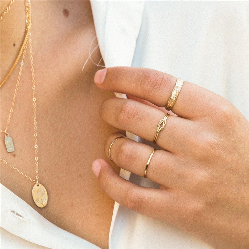 A close-up photo of a woman's hand wearing minimal gold rings on each finger, slightly touching her crisp white linen blouse at the collar, and wearing 4 layered gold necklaces